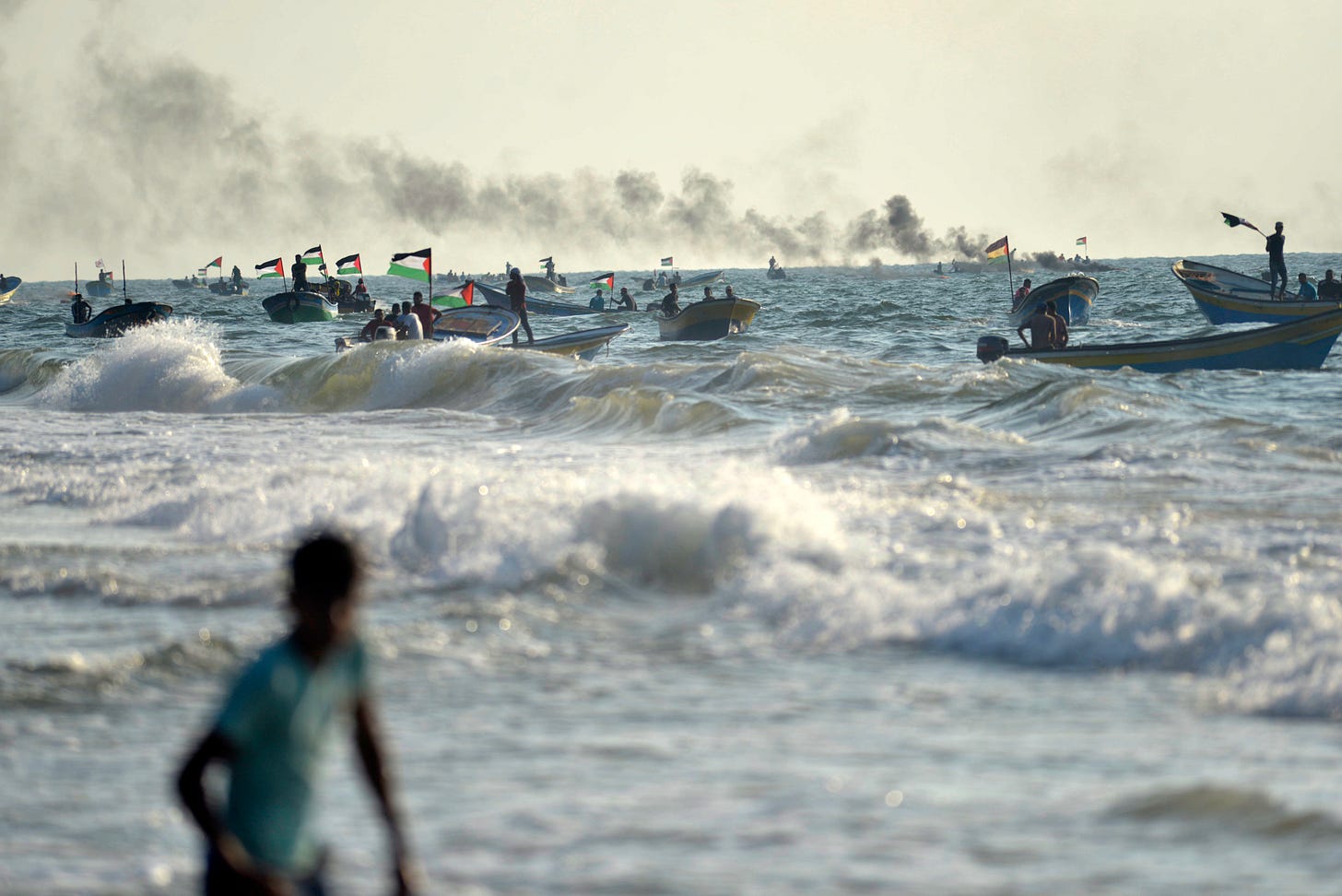 Dozens of Palestinians in small fishing boats fly Palestinian flags on choppy seas off Gaza's coast, with smoke rising in the distance. In the foreground, a young person stands waist-deep in the surf, watching the boats. Dozens of Palestinians in small fishing boats fly Palestinian flags on choppy seas off Gaza's coast, with smoke rising in the distance. In the foreground, a young person stands waist-deep in the surf, watching the boats.