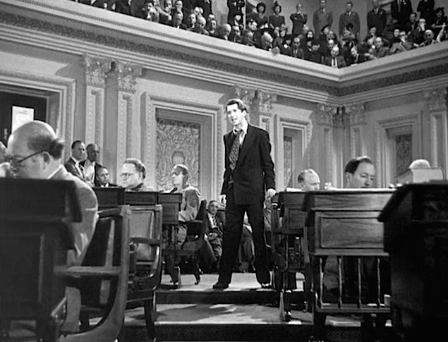 Low-angle black-and-white film still of Jimmy Stewart as Jefferson Smith in Mr. Smith Goes to Washington, bracing himself against a Senate desk during the climactic filibuster, capturing exhaustion and defiance.