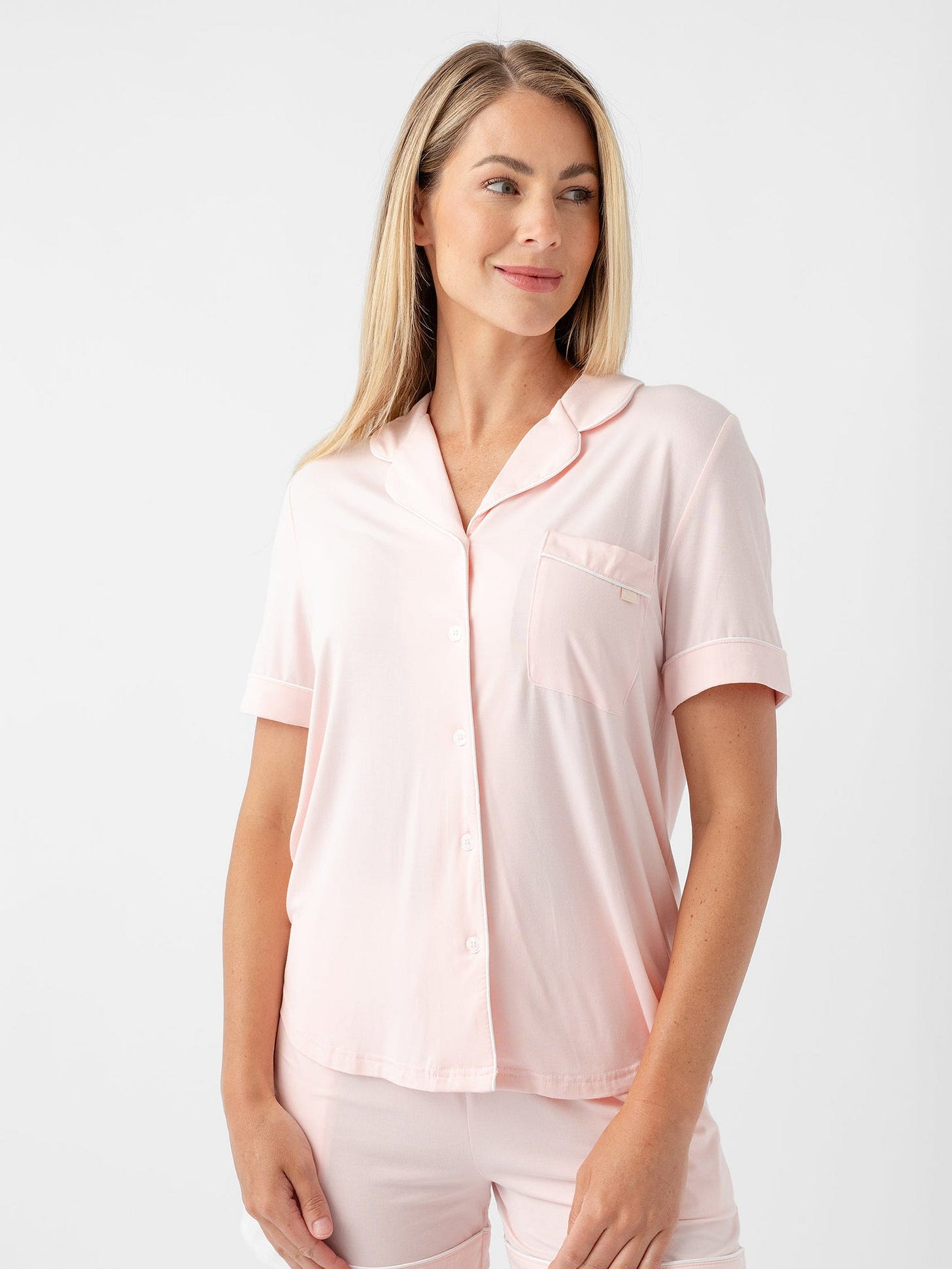 A person with long blonde hair is wearing HIDE's Women's Short Sleeve Bamboo Pajama Top in Stretch-Knit, in light pink, paired with matching shorts. The person is standing and looking to their left, smiling slightly against a plain white background. 