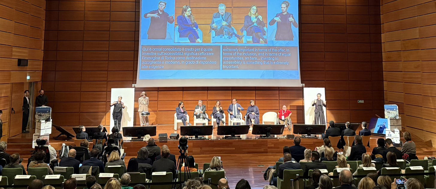 A panel discussion at the World Summit on Accessible Tourism, held in a large auditorium with wooden walls. Seven speakers sit on stage, including a wheelchair user, while two sign language interpreters stand on either side. Above them, a screen displays live captions in Italian and English discussing investment in accessibility. The audience is seated and attentively watching the presentation.