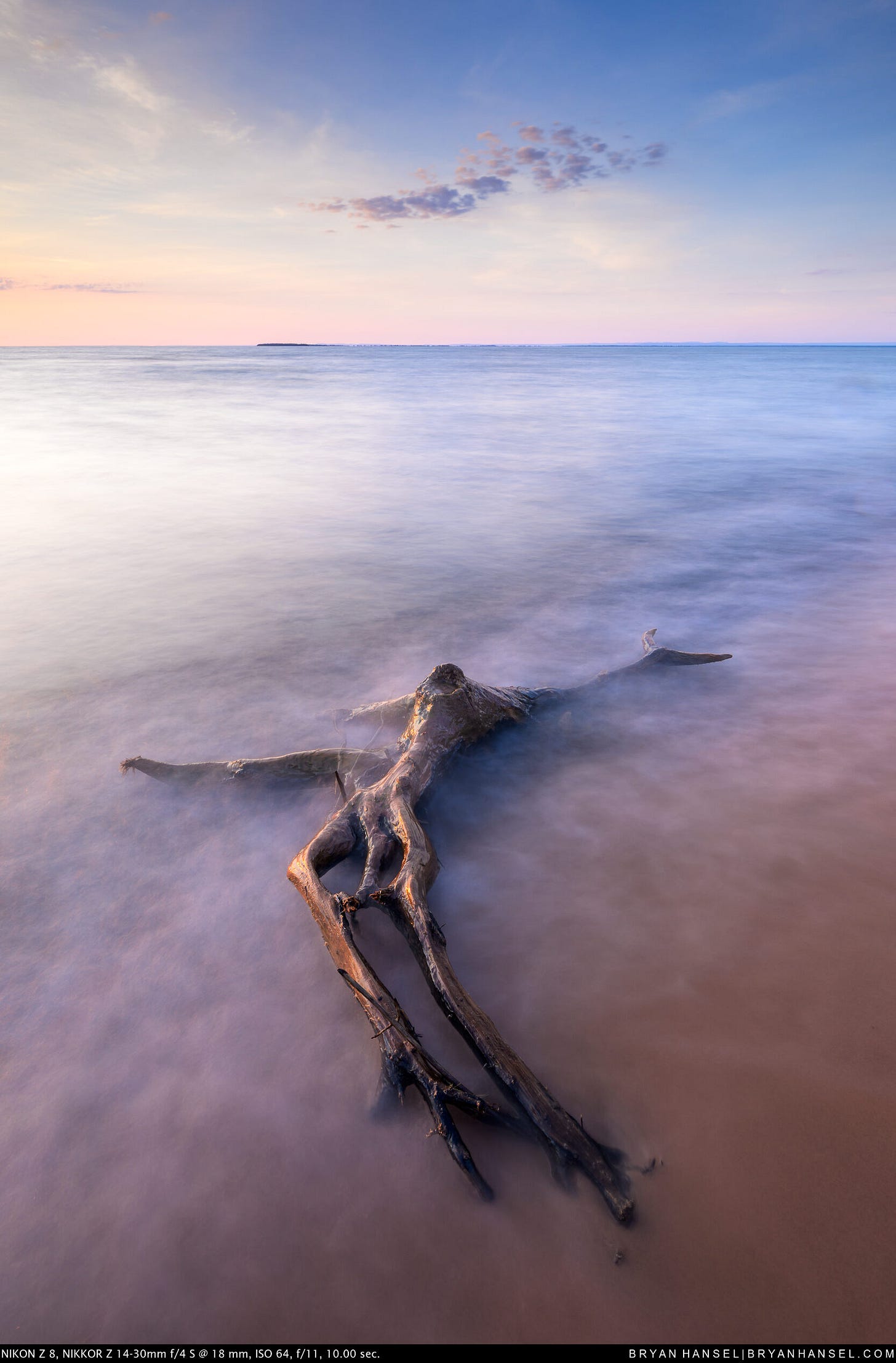A large branching driftwood tree trunk lies across a sandy Lake Superior beach, surrounded by silky long-exposure waves under a calm post-sunset sky with a single small cloud cluster and a distant island on the horizon.