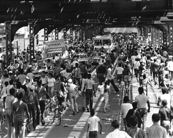 A black-and-white photo of many people walking among cars on the street under an elevated rail line. A black-and-white photo of many people walking among cars on the street under an elevated rail line.