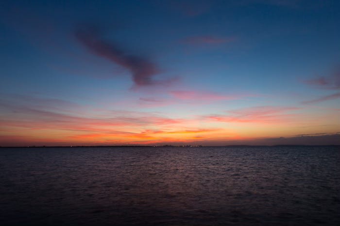 A serene view of Tampa Bay at sunrise. The sky is painted with a gradient of colors, transitioning from deep blue at the top to warm orange and pink hues near the horizon. The sun is just beginning to rise, casting a soft, glowing light across the water. The bay's surface is calm, with gentle ripples reflecting the colors of the sky. In the distance, a faint line of city lights is visible on the horizon, adding a subtle contrast to the natural beauty of the scene.