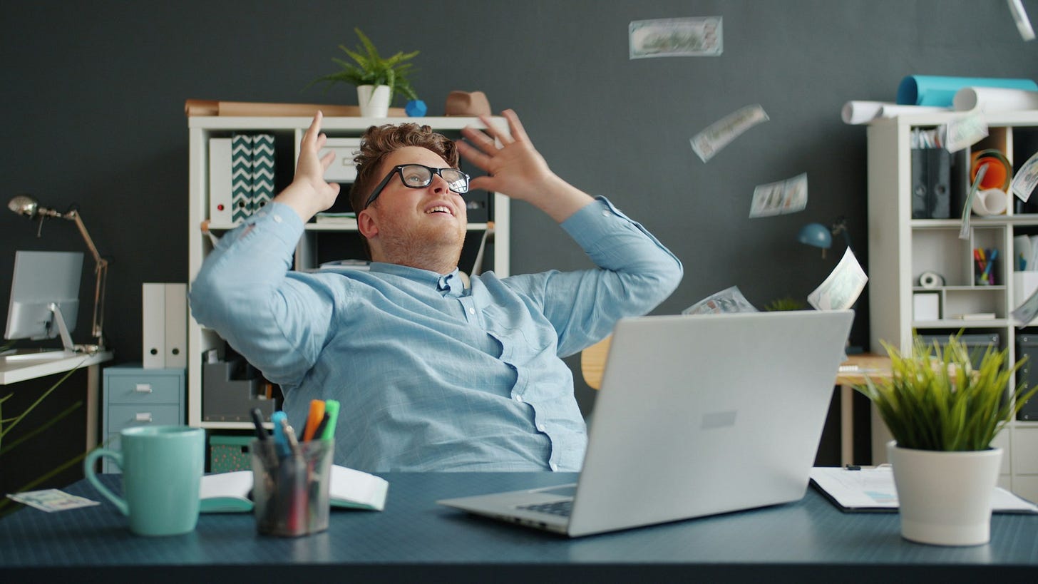 A young man sitting at a desk with a laptop, throwing his hands up in excitement as dollar bills fly around him. Photo by Vitaly Gariev on Unsplash