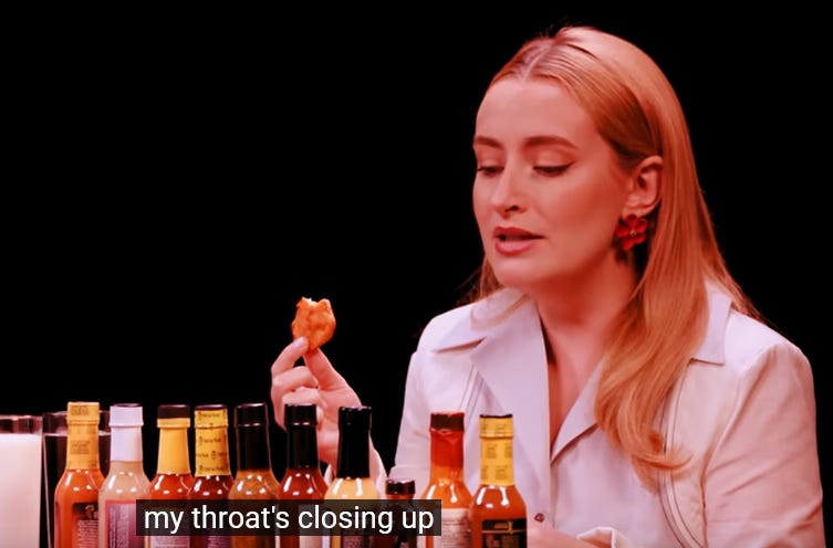 Imagem dos ombros pra cima de Amelia, a mulher loira e branca da imagem anterior, com uma asinha de frango na mão. Existe uma fila de garrafas de molho de pimenta em sua frente. Ela está olhando para o frango mordido chateada. A imagem foi editada para que ela fique ligeiramente vermelhada. O fundo é preto. A legenda de sua fala: my throat's closing up