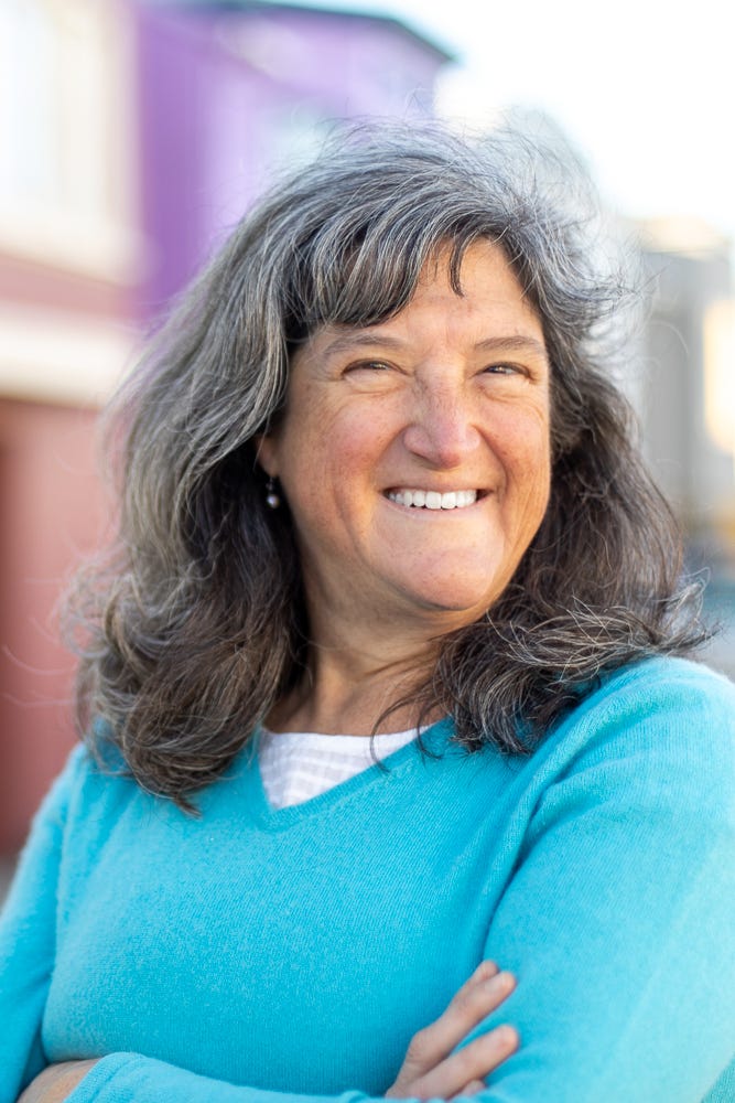 A head shot of Mira, a Gen X white woman with salt-and-pepper, shoulder-length hair. She's smiling a confident smile. She's wearing a turquoise sweater and white shirt and has her arms folded across her chest. 
