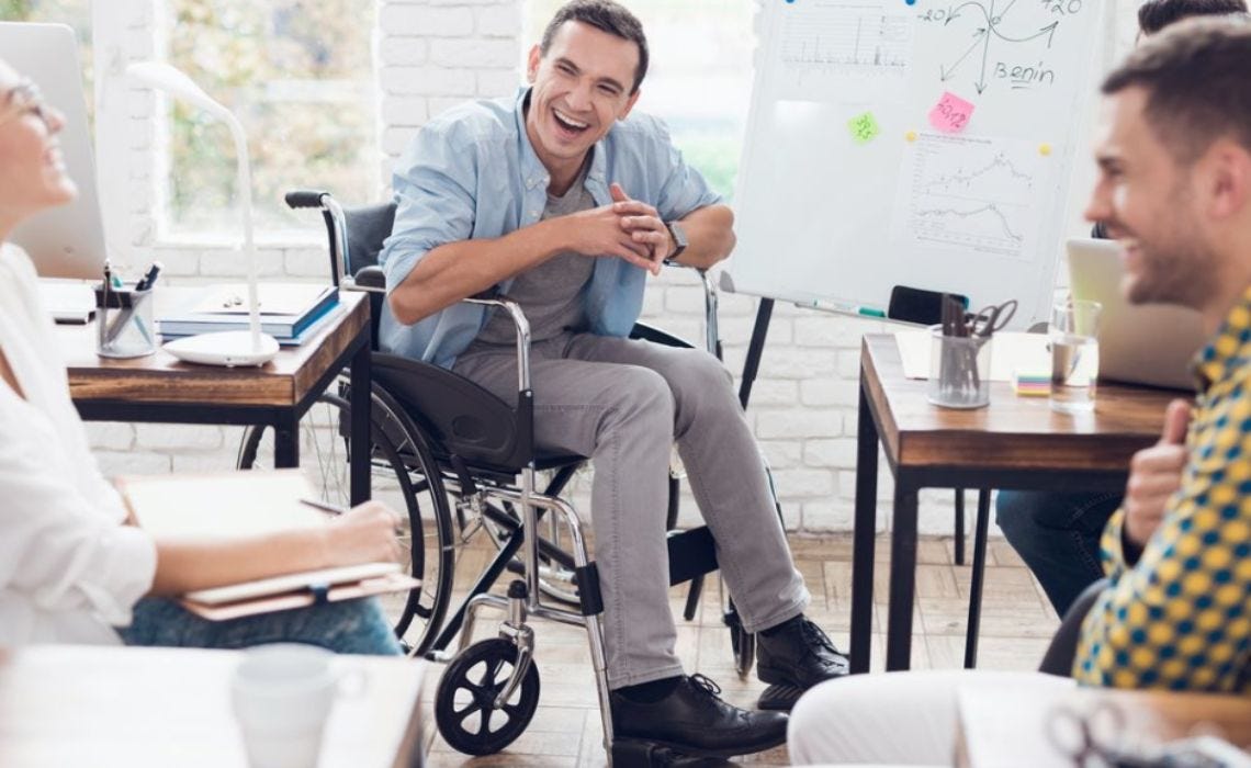 A man in a casual shirt and jeans sits in a wheelchair, laughing with colleagues during an office meeting. They are seated at desks with notebooks and laptops, and a whiteboard with charts and notes is in the background.