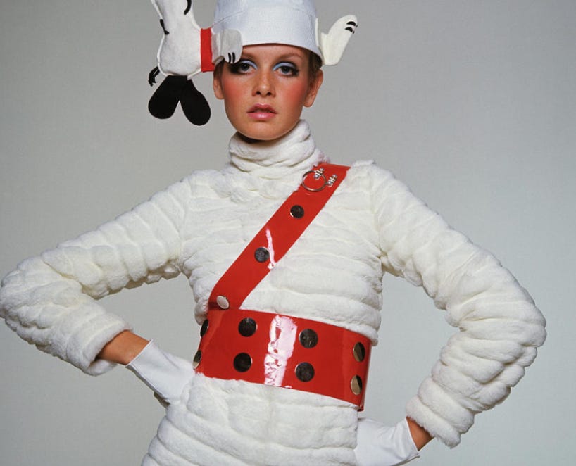 A 1967 photo of British model Twiggy, taken by Burt Stern for Vogue. Twiggy is facing the camera head-on, with long lashes and light blue eye makeup, pink blush, and her mouth slightly open. She is wearing a hat featuring Snoopy sleeping sideways, a white puffy coat, and a red plastic belt across her waist and chest. She is wearing short white gloves and has her hands on her hips.