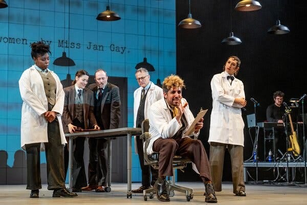 Actors in lab coats and suits strike poses during a scene from a play.