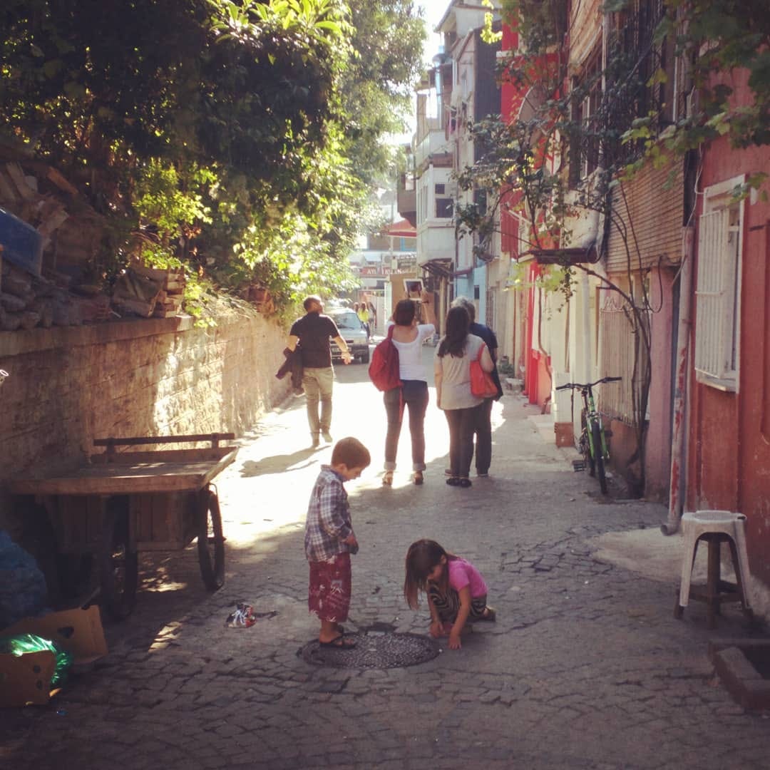 Sevimli dar bir sokak, sokak bile denemez o kadar dar. Solda insan boyuna yakın bir taş duvar ve üzerinden taşmış ağaçlar, bitkiler. Sağda sevimli, apartmanlar. Ortada iki çocuk, 4-5 yaşlarında, bir kız, bir oğlan, ikisi de bir mazgala bakıyor. Oğlan ayakta, kız yere çömelmiş. Kızın saçları sarkmış. Çocukların arkasında 4 yetişkin, iki erkek iki kadın, etrafa bakarak yürüyorlar. Çocukların olduğu yer gölge, yetişkinlerden itibaren parlak bir sonbahar ışığı.
