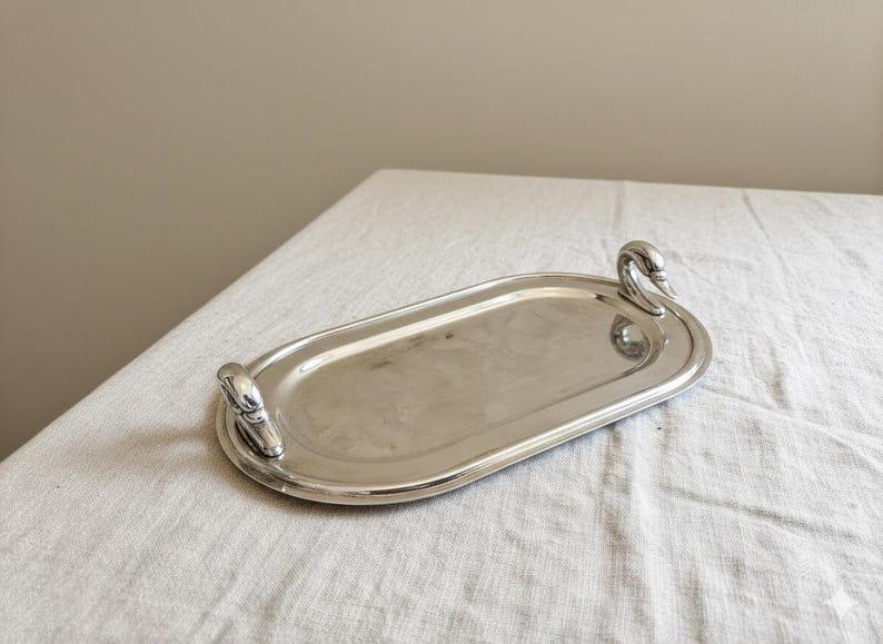 May include: An oval-shaped silver-toned metal serving tray featuring two handles, each sculpted in the form of a swan head. The tray has a smooth, reflective surface and a raised rim, providing a polished appearance for this functional serving piece.