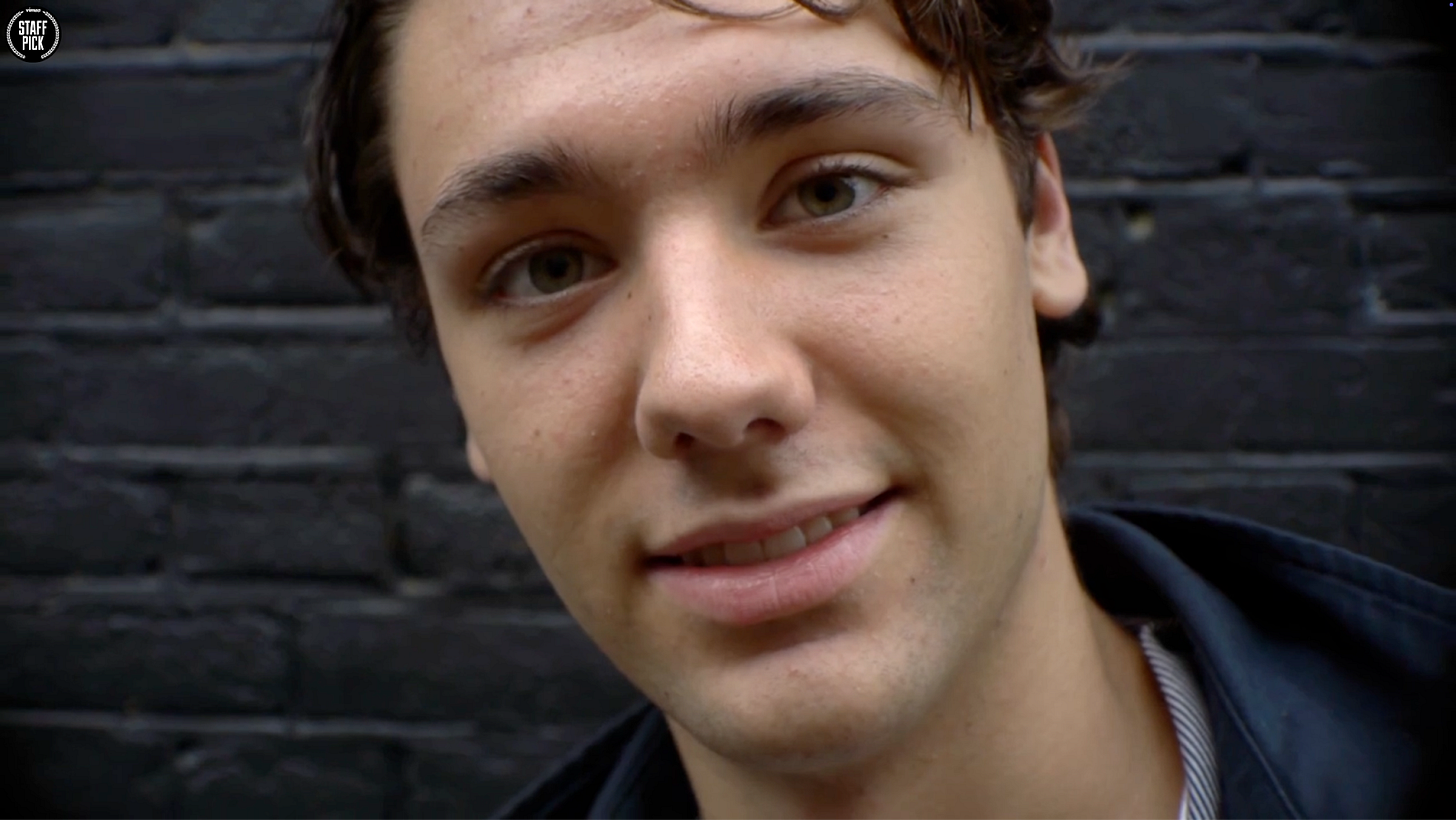 Close-up portrait of a white teenage boy with brown hair wearing a dark jacket, smiling slightly at the camera against a brick wall background.