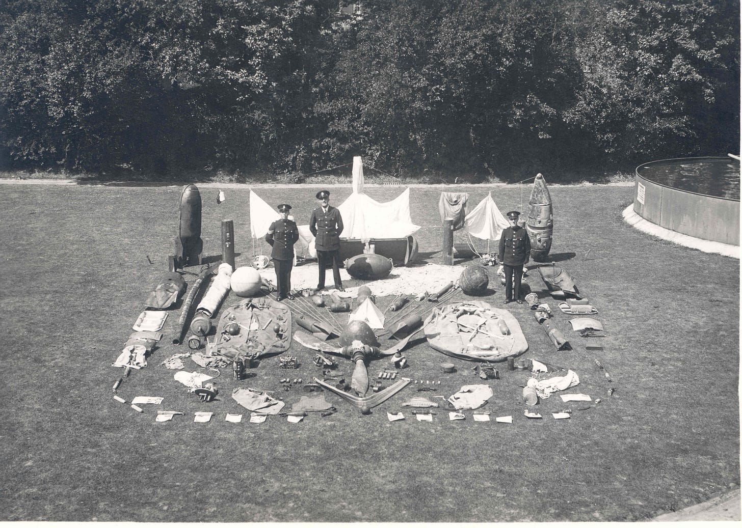 Three uniformed police officers stand amongst arranged debris and shrapnel, ranging from tiny 1ft-long incendiary bombs to larger bombs around the height of the officers. Some are accompanied by parachutes while others have been reduced to parts. The central feature is a large bent up propellor.
