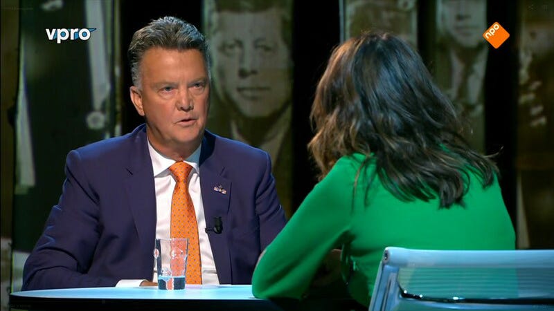 Screenshot van de aflevering van zomergasten waar Louis van Gaal te gast is. Je ziet Louis aan tafel zitten in een blauw pak en oranje stropdas, Janine zie je van achteren