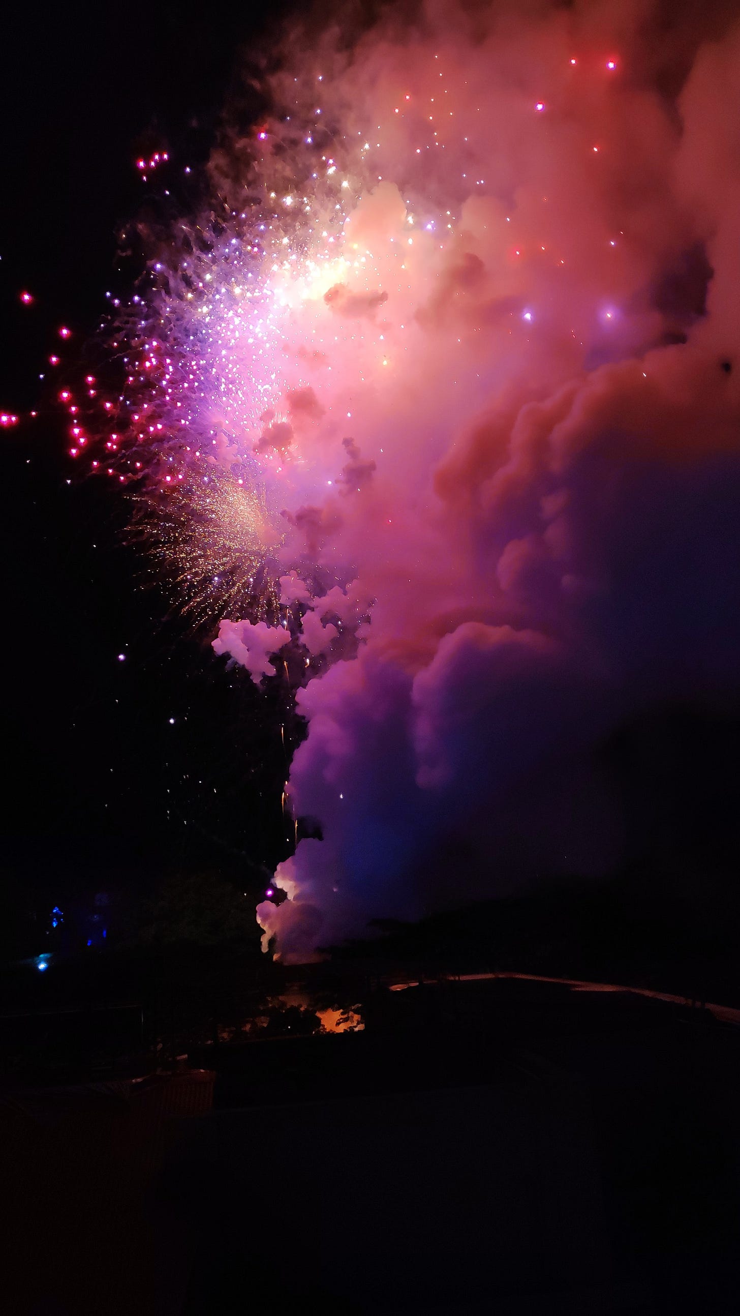 Fireworks going off. Magenta and purple lots of smoke fillng the right side of the frame, glowing to match the color of the explosions.