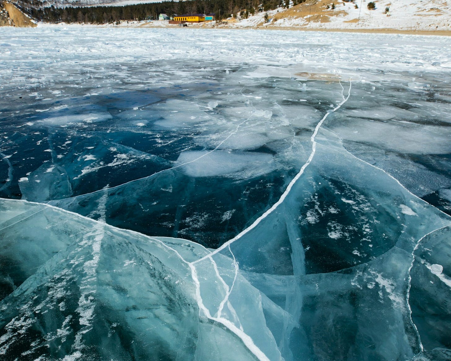A frozen lake with deep blue ice and long white cracks running across the surface, revealing hidden webs of fracture beneath.