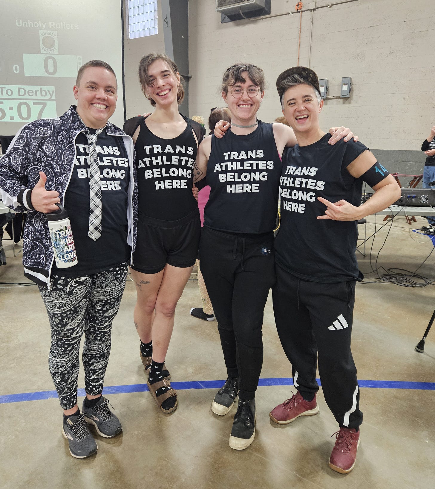 Four humans stand, arm-in-arm, smiling and proudly displaying shirts that read "TRANS ATHLETES BELONG HERE." They're all cute and trans and/or non-binary.