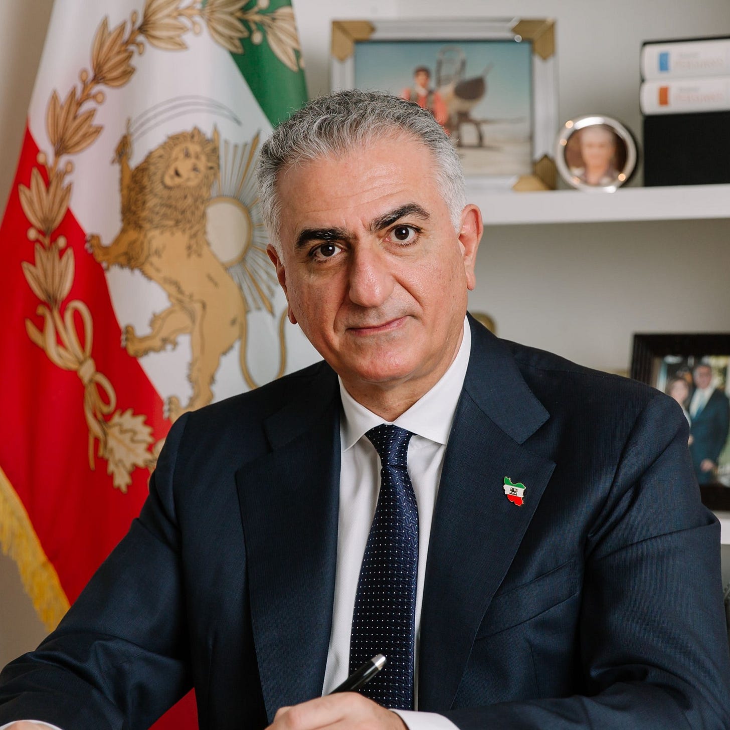 Reza Pahlavi sitting in front of the flag of the Iranian monarchy, featuring the Lion and Sun.