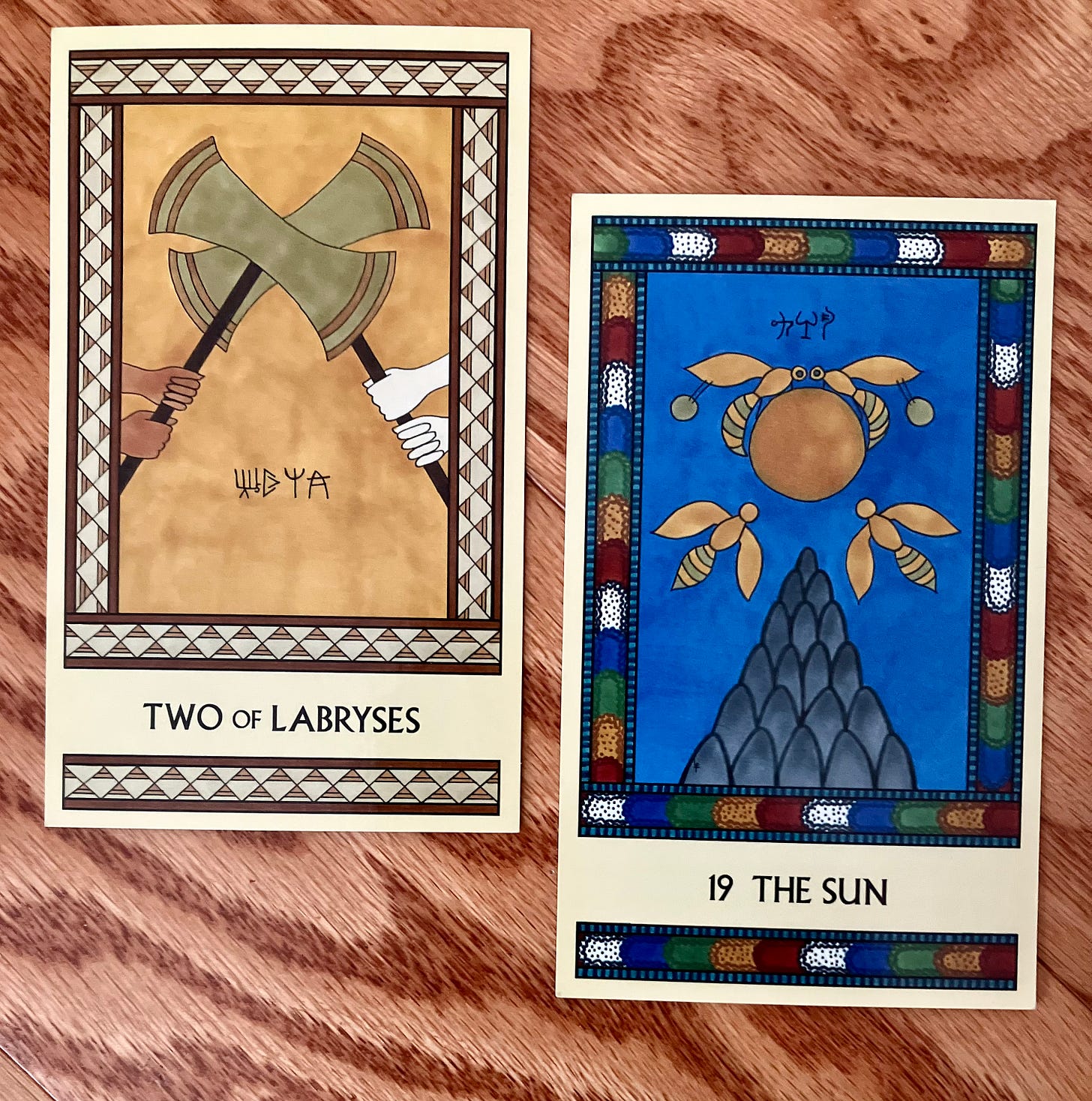 Two Minoan Tarot cards side by side on a wood surface. The Two of Labryses is in shades of gold and cream. It shows two pairs of hands each holding a large labrys, the two labryses locked together so neither can win or leave. The Sun has a multicolor border and shows several bees circling the Sun above a tall mountain with a deep blue sky in the background.