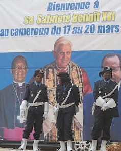 Female soldiers stand in front of a billboard featuring three men, two of them in religious garb. Female soldiers stand in front of a billboard featuring three men, two of them in religious garb.