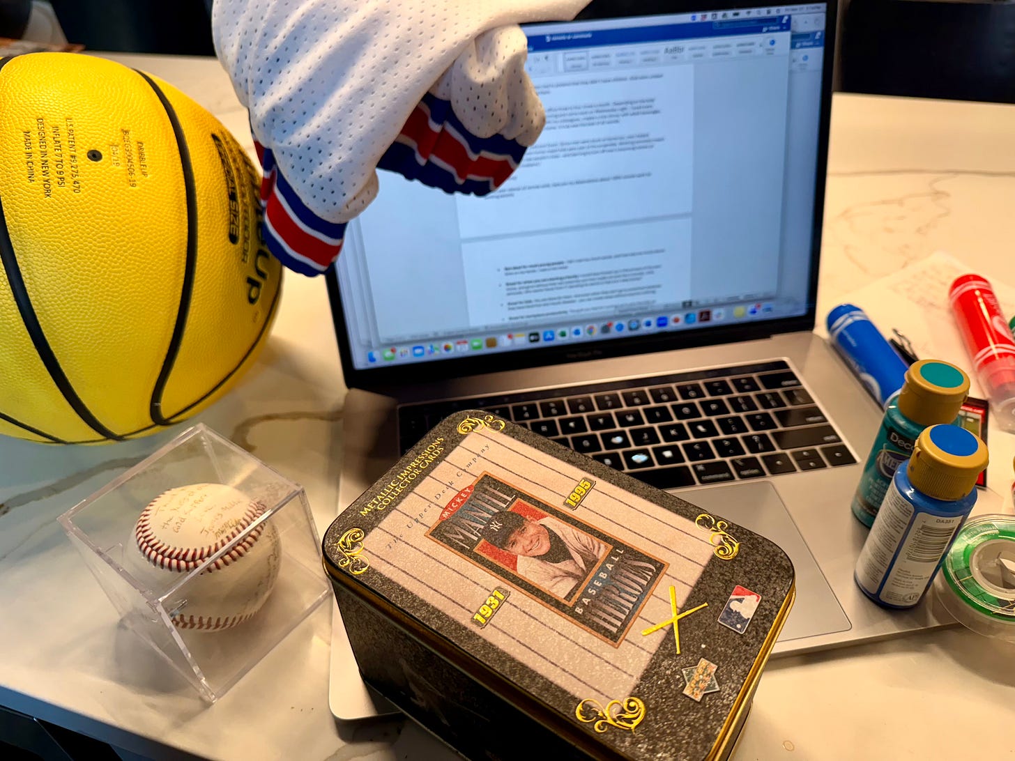 Photo of a laptop surrounded by clothes, markers, paints, basketball and baseball depicting work from home chaos.