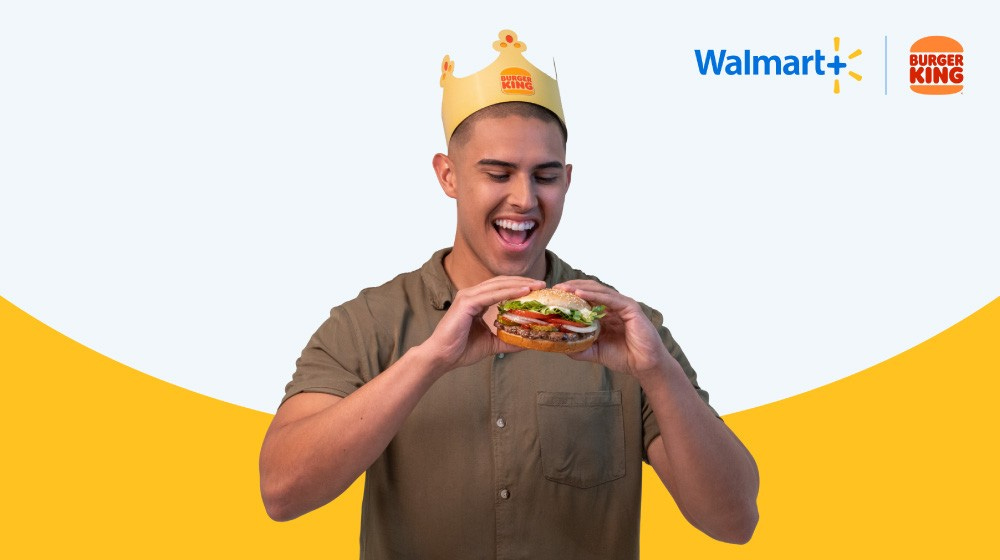 A man in a brown button down shirt wears a Burger King crown and with both hands is holding a Whopper hamburger. The man is looking at the burger and smiling. The Walmart Plus and Burger King logos are in the top right corner.