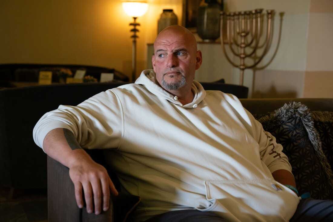 Senator John Fetterman sits in a room in Jerusalem with a menorah in the background. Fetterman is wearing a white hooded sweatshirt and looking off camera. 