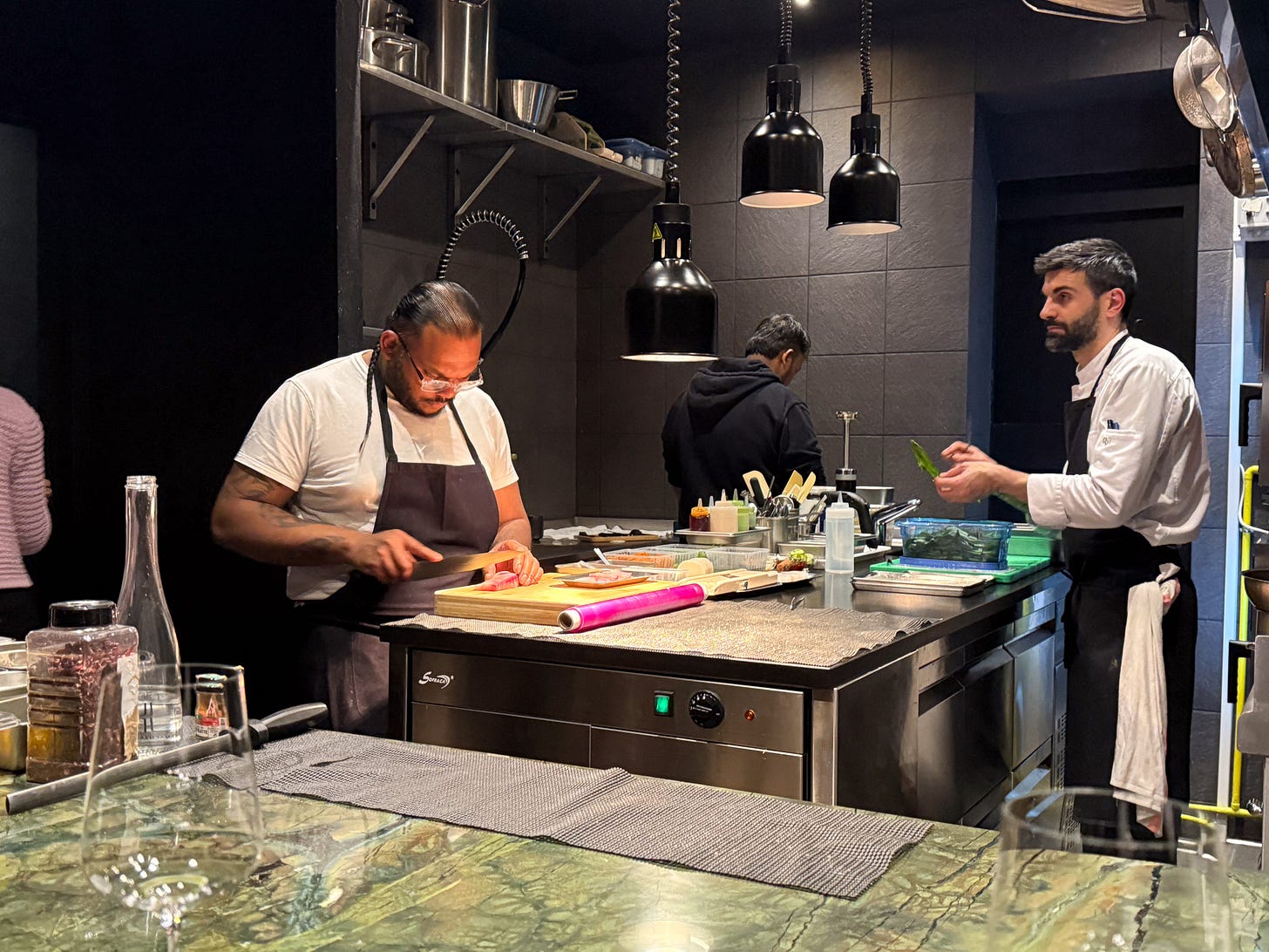 The intimate dining room at Mantra restaurant showing dark walls, counter seating facing the open kitchen with marble-topped counter, and diners at round tables