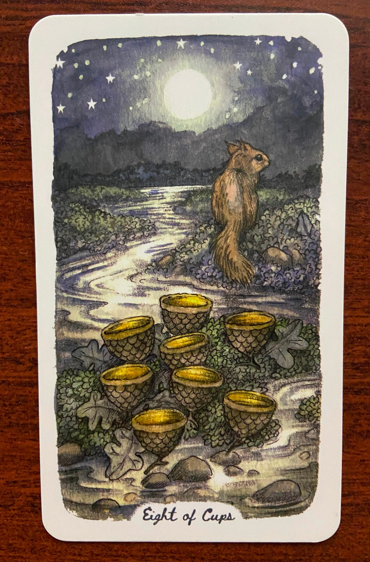 Top card, Four of Wands: Two sweet-faced foxes sit side by side in a field of wildflowers. Their head are crowned by leaves. Behind them stand four slender flowering trees, and a few butterflies are aloft in the air.  Bottom card, Eight of Cups: Under a blazing full moon and a sky of stars, a river flows. A squirrel sits on land facing dark mountains in the distance. Eight “cups”--empty acorns, surrounded by oak leaves–rest in the foreground of the image. The squirrel has left them behind.