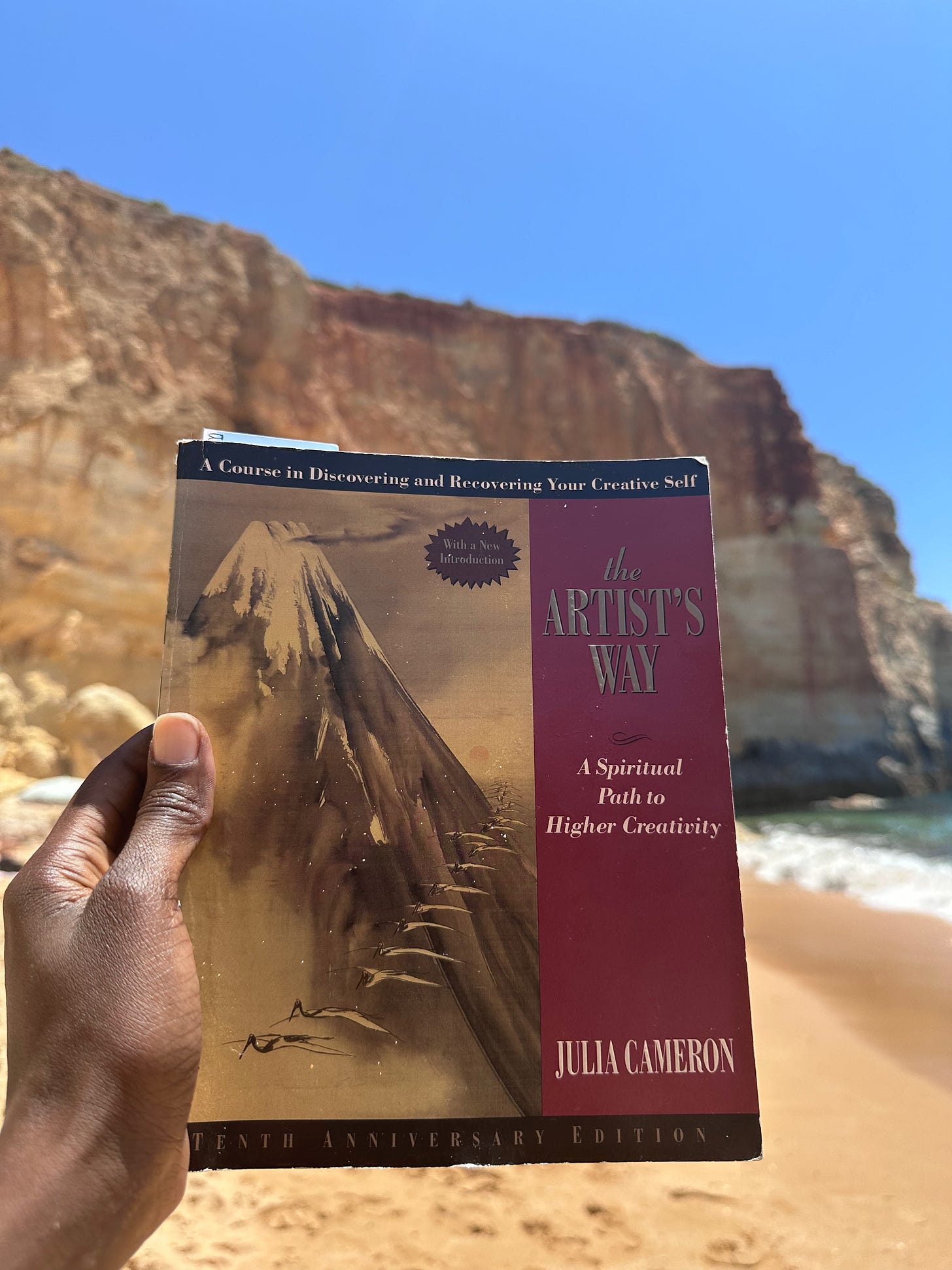 The Artist's Way by Julia Cameron, held on a sandy beach in front of tall sea cliffs" Caption: "The book that got me writing again.