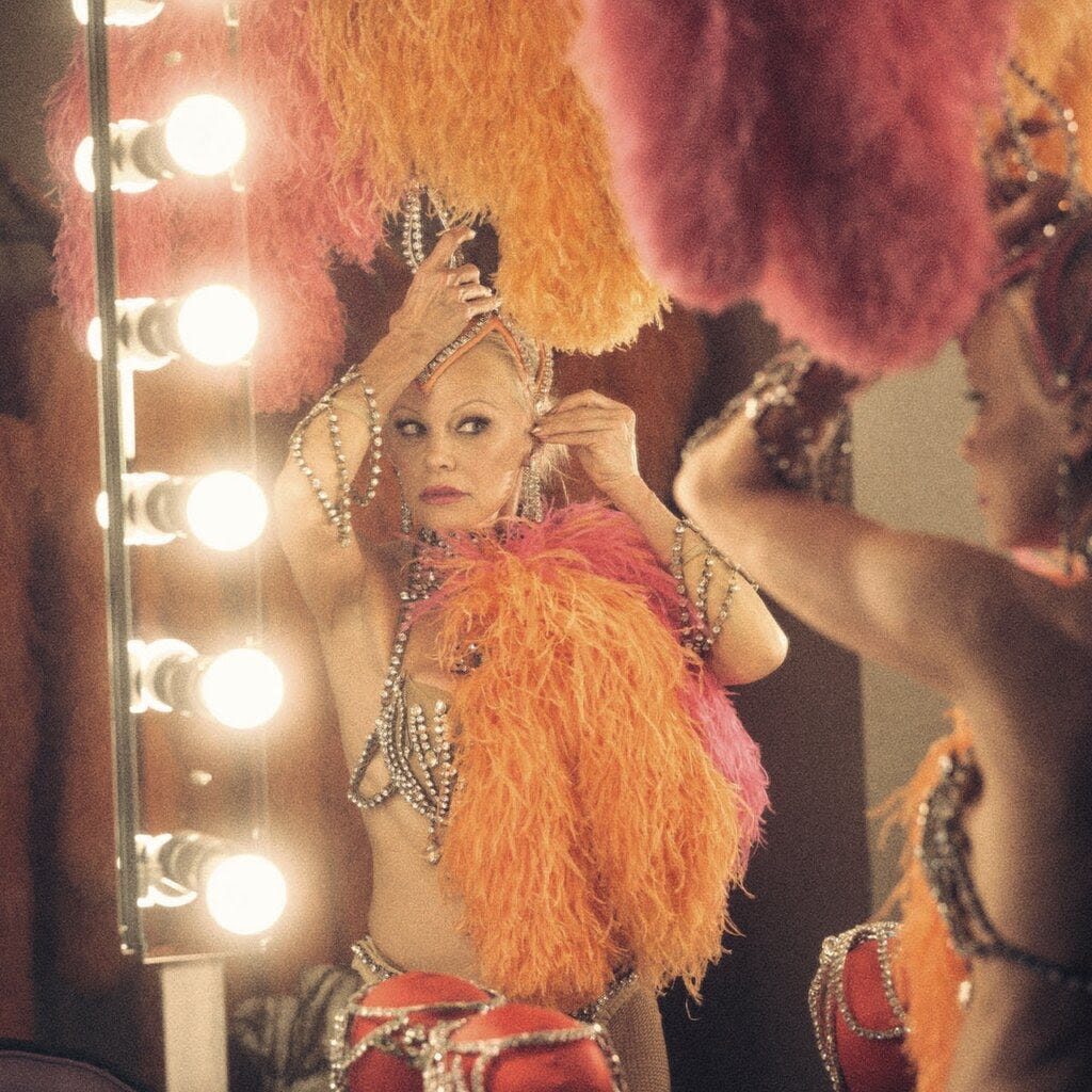 Pamela Anderson, in a skimpy, feathery showgirl costume, adjusting her headpiece in a vanity mirror.