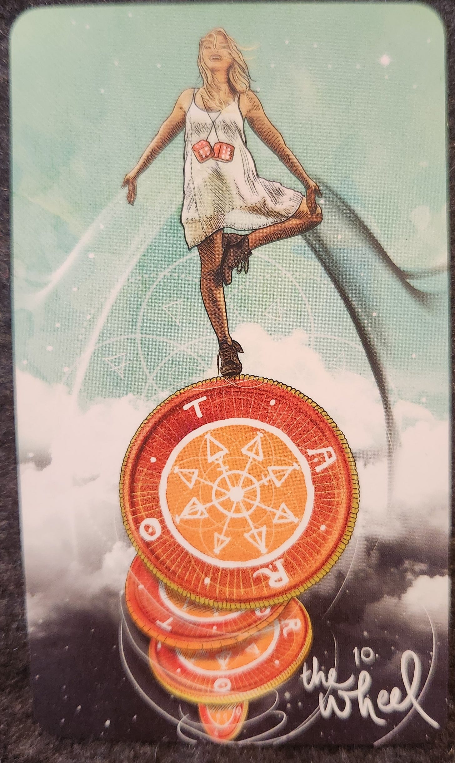A tarot card labelled 10. The Wheel. A woman stands balanced on the edge of a large wheel, covered in arrows pointing from the centre of the wheel to the edges. Around the edges of the wheel, spaced at each quarter, are the letters “T”, “A”, “R”, and “O”. The woman is standing in tree pose, standing on one leg with the other bent and the feet pressed around her inner thigh. Her eyes are closed, her arms are outstretched. She looks calm. Around her neck she is wearing a pair of red dice.