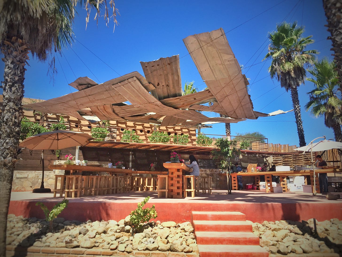 Outdoor bar and dining area at Conchas de Piedra restaurant in Valle de Guadalupe, showing a sculpted wooden roof structure over wooden bar stools and tables, framed by palm trees. Outdoor bar and dining area at Conchas de Piedra restaurant in Valle de Guadalupe, showing a sculpted wooden roof structure over wooden bar stools and tables, framed by palm trees.