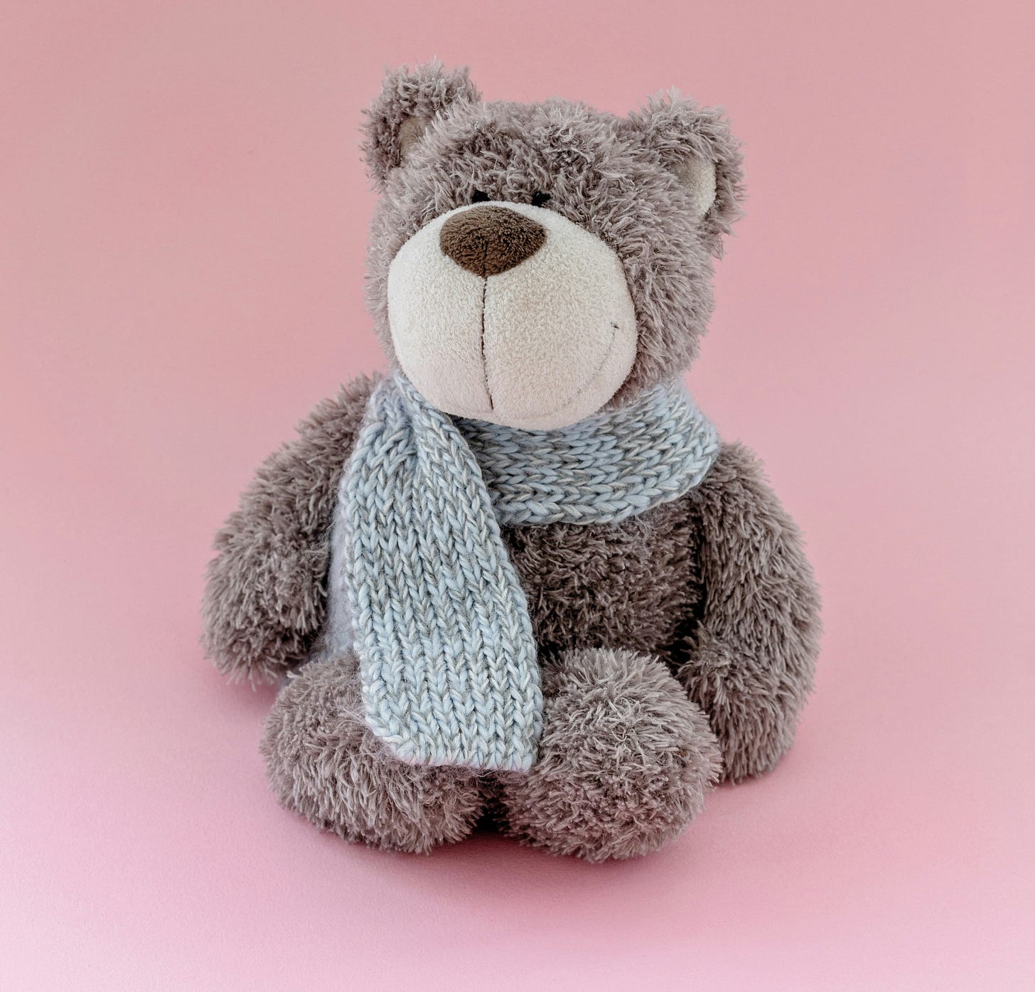Smiling soft light brown teddy bear in a light grey scarf, against a light pink background Smiling soft light brown teddy bear in a light grey scarf, against a light pink background
