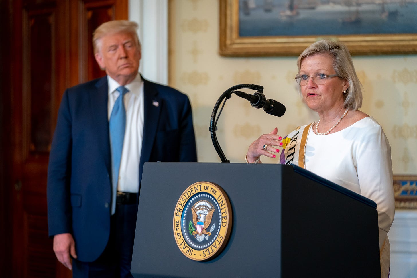 Cleta Mitchell speaking at a lectern during a 19th Amendment centennial event at the White House, with then-President Donald Trump standing in the background near the presidential seal. Cleta Mitchell speaking at a lectern during a 19th Amendment centennial event at the White House, with then-President Donald Trump standing in the background near the presidential seal.