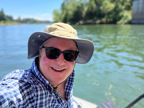Portland Rose banner, Mt Hood, man wearing a hat, sunglasses, and blue shirt by the water