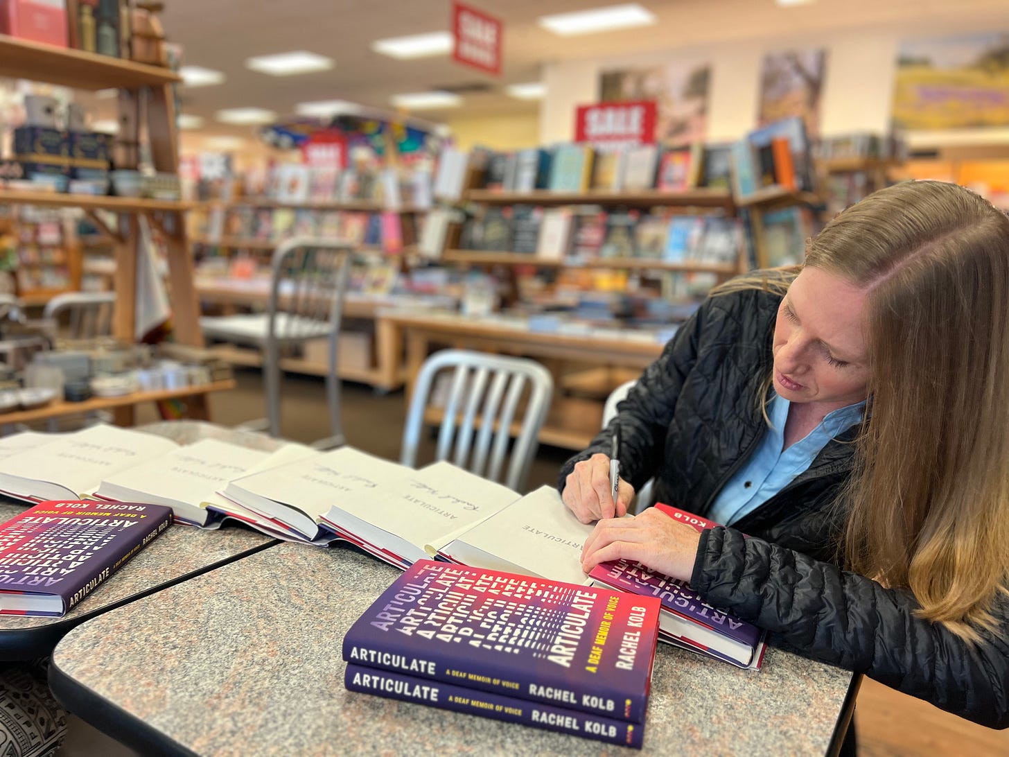 I'm sitting at a table in a black jacket, signing several copies of ARTICULATE spread out in front of me. A stack of even more books is in the foreground.