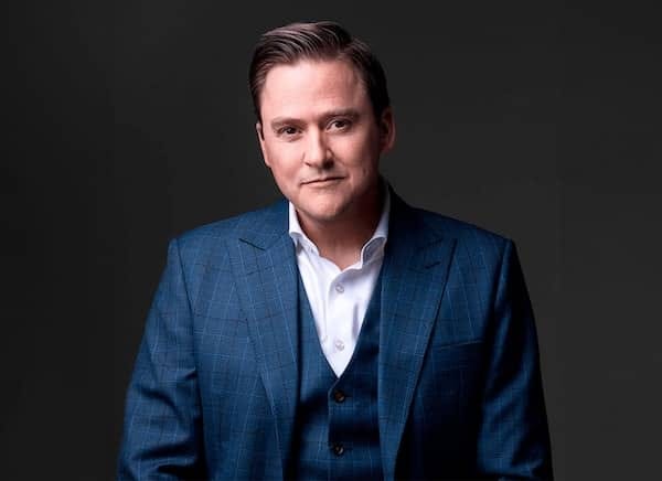 A man in a blue suit and white shirt poses confidently against a dark background.