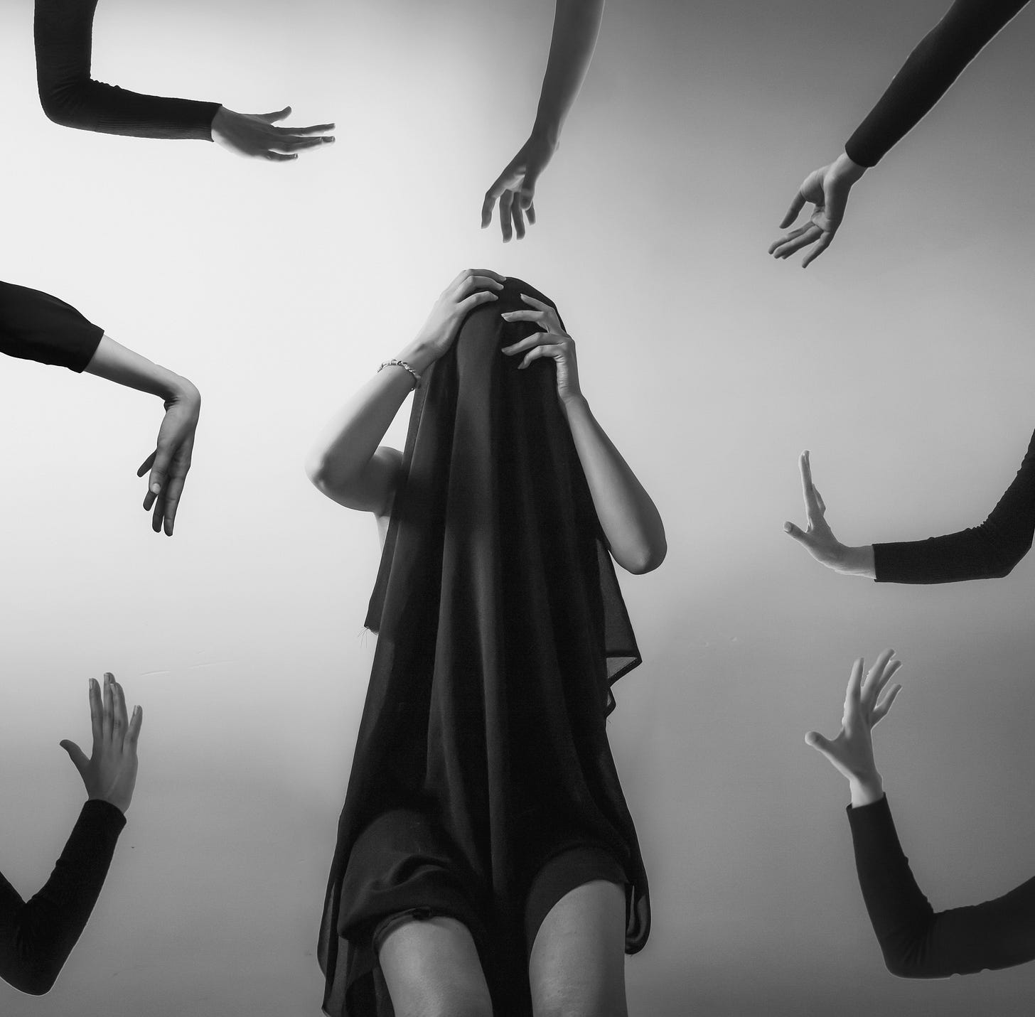 A woman sits, covered by a black fabric while multiple arms reach around her.