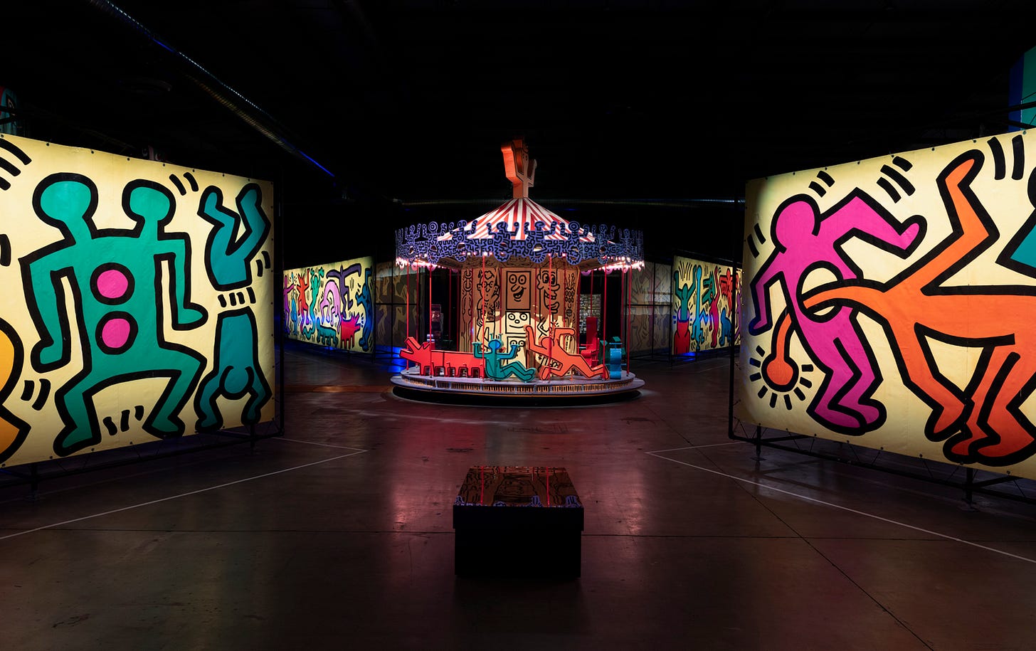 An overview of a reassembled, historic amusement park inside a warehouse. The image focuses on a colorful carousel in the center, designed by artist Keith Harnig, with illuminated walls also covered in his cartoonish drawings.