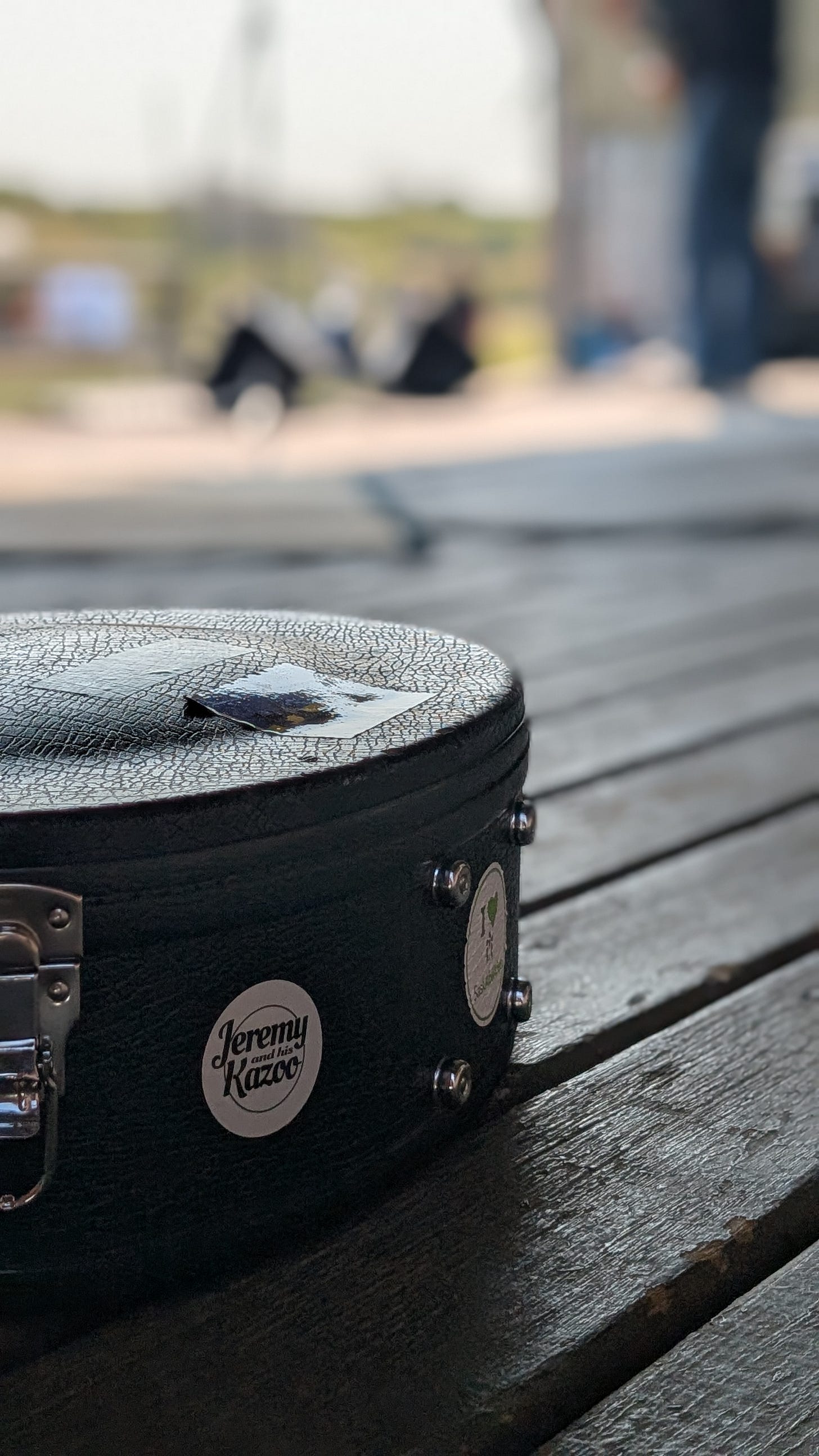 A round silver "Jeremy and his Kazoo" sticker on a guitar case set on a stage. 