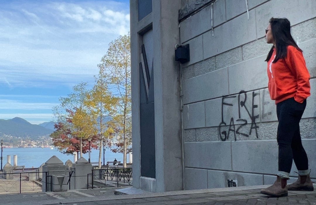 Justine in an orange hoodie standing next to graffiti that reads: FREE GAZA. In the background are the mountains and Pacific Ocean.