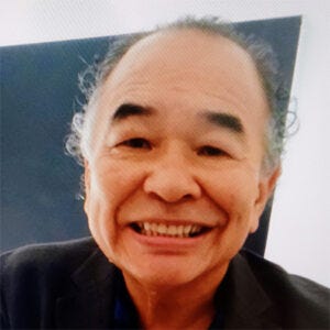 Homem idoso, de cabelos grisalhos e curtos, aparece sorrindo em um retrato. Ele veste uma camisa azul-escura sob um blazer preto e está sentado diante de uma parede de fundo escuro. Homem idoso, de cabelos grisalhos e curtos, aparece sorrindo em um retrato. Ele veste uma camisa azul-escura sob um blazer preto e está sentado diante de uma parede de fundo escuro.