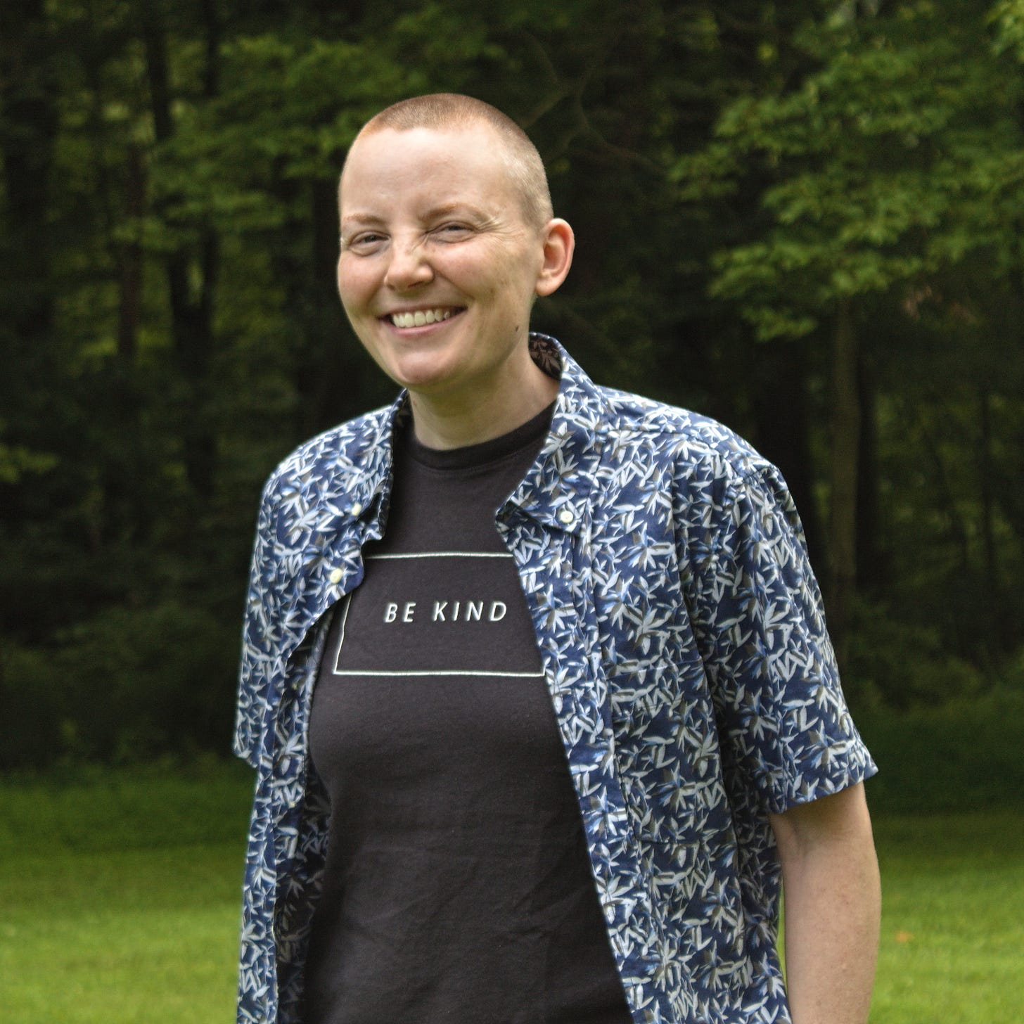 Ryn, white person smiling at camera in front of some trees, wearing a t-shirt that says "Be Kind" Ryn, white person smiling at camera in front of some trees, wearing a t-shirt that says "Be Kind"