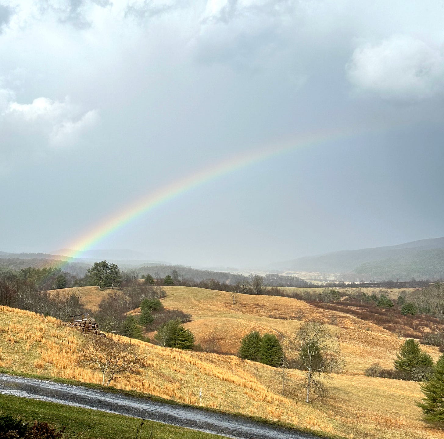 A photo of some rolling hills with storm clouds above them and a rainbow shooting through the sky