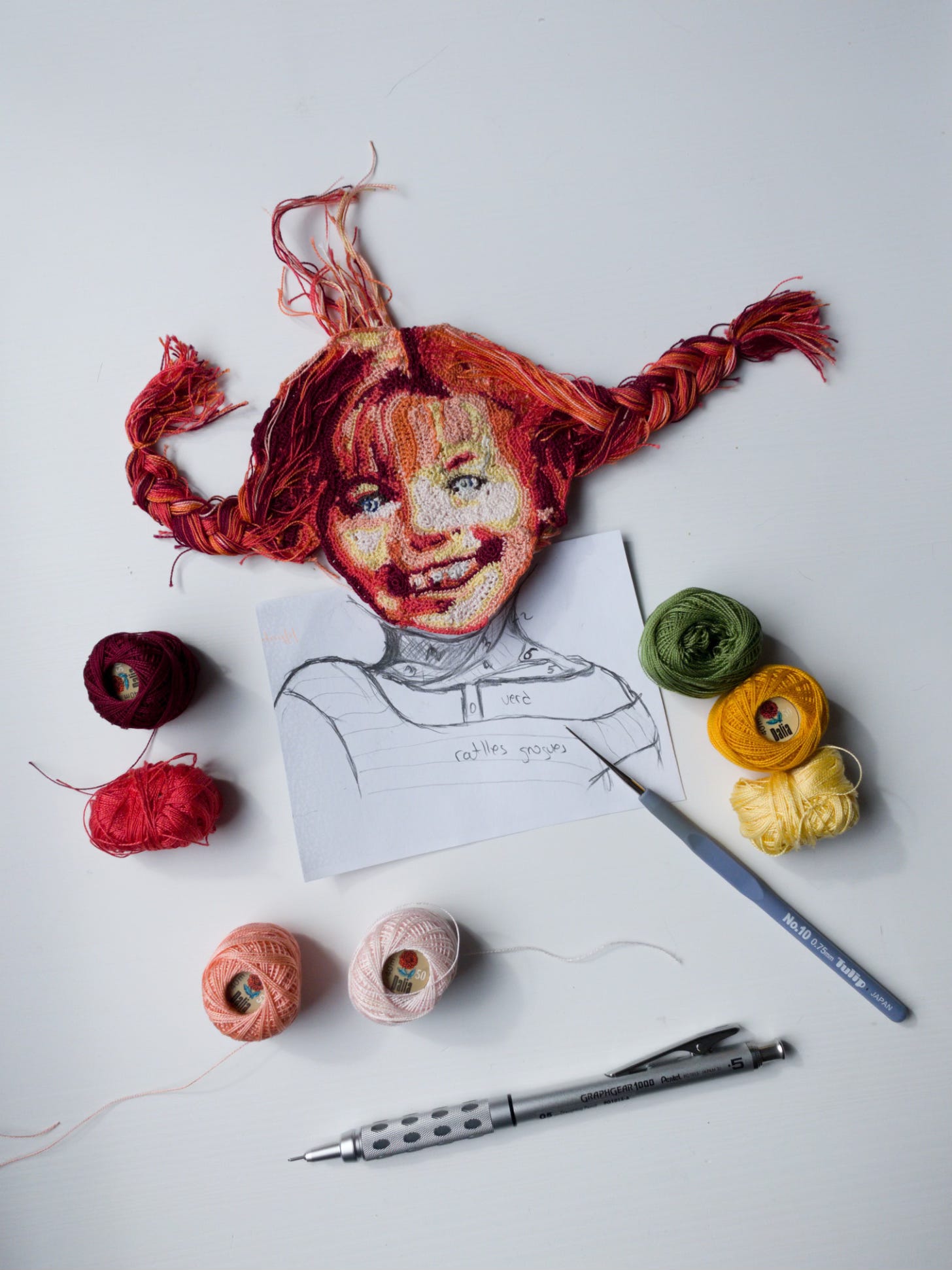 Crochet portrait of Pippi Langstrumpft lays on a white table. There is a pencil sketch of her body to finish the piece. There are also threads, a crochet hook and a mechanical pencil.