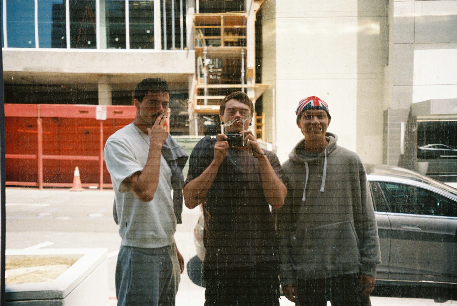 Three young men taking a photo in a window reflection on 35mm film. Dressed casually. The left smoking a cigarette, the middle holding the camera, and the right smiling wearing a Union Jack beanie hat. The glass reflecting them is dirty.