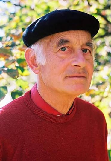 A photo of a grey haired man wearing a black beret and a red jumper. There are trees in the background.