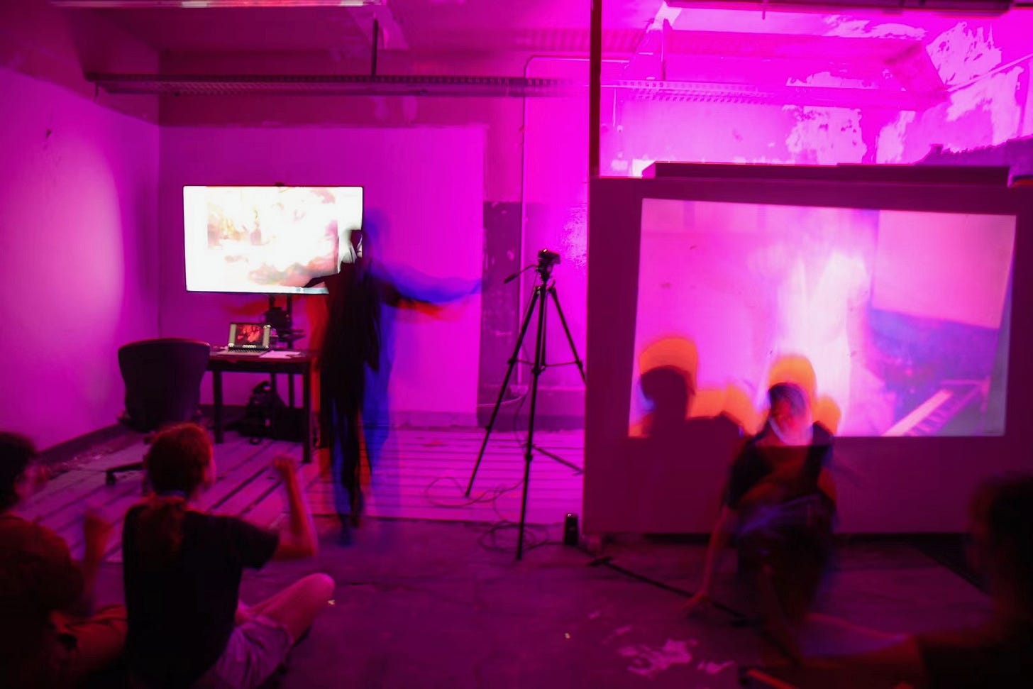 Performance still: A room illuminated by intense magenta lighting. A performer in dark clothing moves in front of a projection screen, creating a blurred effect. Audience members sit on the floor, reacting to the visuals and movement. A camera on a tripod suggests a live recording or interactive media element.