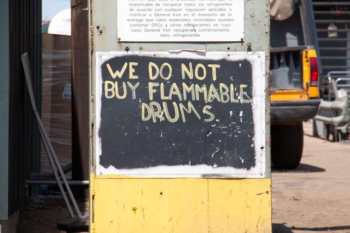 A hand-painted sign reading "WE DO NOT BUY FLAMMABLE DRUMS" on a metal surface, with part of a yellow pickup truck in the background at a Chicago scrap yard. A hand-painted sign reading "WE DO NOT BUY FLAMMABLE DRUMS" on a metal surface, with part of a yellow pickup truck in the background at a Chicago scrap yard.
